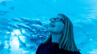 Ein Mädchen mit Brille lächelt begeistert, während sie unter einer blauen Eisdecke steht, die Licht reflektiert.