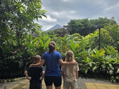Ein Erwachsener steht mit einem Kind und einem weiteren Jugendlichen vor einem grünen Garten, im Hintergrund der Arenal-Vulkan.