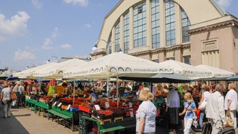 Ein belebter Markt mit bunten Ständen, die frisches Obst und Gemüse unter Sonnenschirmen anbieten, umgeben von vielen Menschen.