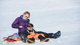 Eine Frau und ein Junge sitzen entspannt auf einer schneebedeckten Fläche, beide tragen Sonnenbrillen und warme Kleidung.