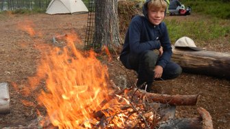 Ein Junge sitzt lächelnd neben einem lodernden Lagerfeuer, umgeben von Bäumen und Zelten in der Natur.