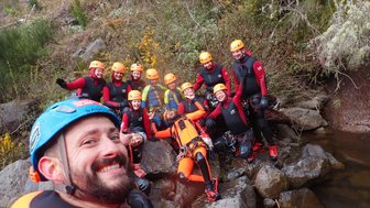Eine Gruppe von zehn Personen in Neoprenanzügen und Helmen posiert fröhlich am Ufer eines Gewässers.