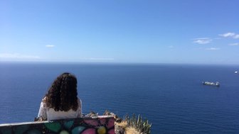 Eine Person mit lockigem Haar sitzt auf einem Felsen und blickt auf das weite, ruhige Meer unter einem strahlend blauen Himmel.