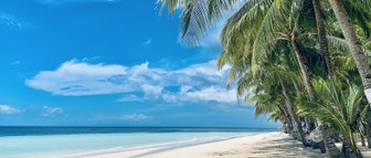 Ein ruhiger Strand mit feinem, weißen Sand erstreckt sich unter einem strahlend blauen Himmel und sanften Wolken.