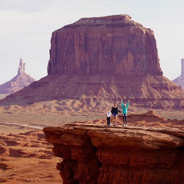 Drei Personen stehen auf einer Felsplattform in Monument Valley, umgeben von beeindruckenden roten Felsen und Hügeln.