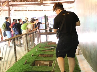 Ein Besucher steht auf einem schmalen Weg über den Cu Chi Tunneln, umgeben von Gras und einer Gruppe von Touristen im Hintergrund.