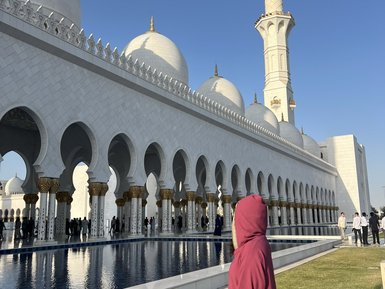 Eine Person in einem roten Gewand steht am Rand eines Wasserspiegels, während die beeindruckende Sheikh Zayed Moschee im Hintergrund sichtbar ist.