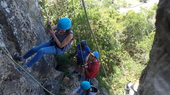 Eine Person mit blauer Kletterhelm schwingt an einem Seil an einer Felswand, während andere Kletterer im Hintergrund stehen.