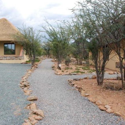 Zwei Hütten mit Strohdächern stehen an einem Kiesweg, umgeben von niedrigen Büschen und einem bewölkten Himmel.
