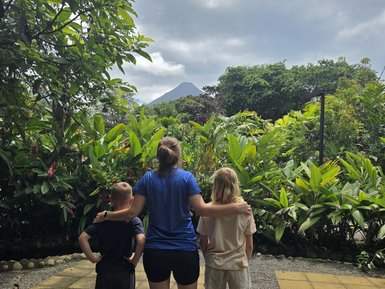 Ein Erwachsener steht mit einem Kind und einem weiteren Jugendlichen vor einem grünen Garten, im Hintergrund der Arenal-Vulkan.