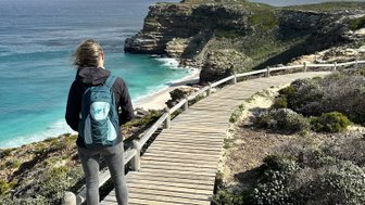 Eine Frau mit einem Rucksack steht auf einem Holzsteg und blickt auf die Küste mit türkisfarbenem Wasser und Klippen.