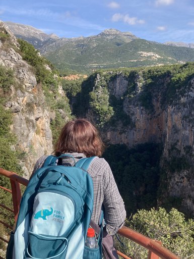 Eine Person mit einem blauen Rucksack steht an einem Geländer und blickt in eine tiefe Schlucht, umgeben von Bergen.