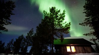 Ein Holzhaus steht in einer verschneiten Landschaft, während die Nordlichter in leuchtendem Grün darüber tanzen.