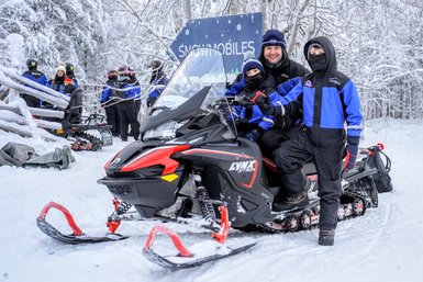 Eine Familie sitzt auf einem Schneemobil in einer verschneiten Winterlandschaft, umgeben von schneebedeckten Bäumen.