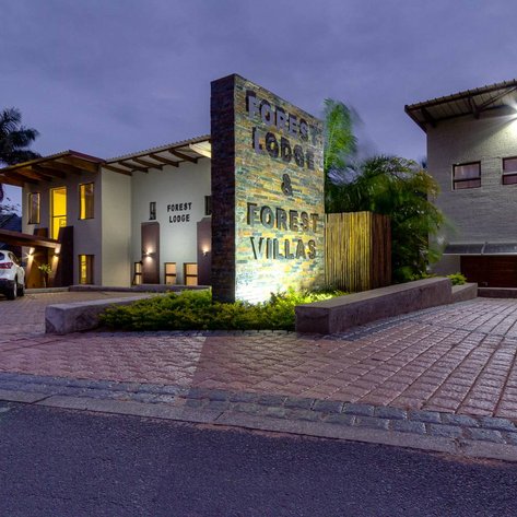 Ein modernes Gebäude mit dem Schild 'Forest Lodge' und 'Forest Villas' vor einer gepflasterten Einfahrt bei Dämmerung.