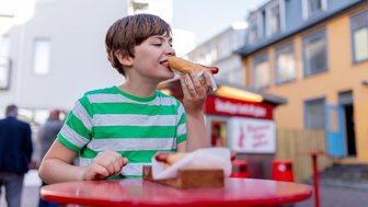 Ein Junge mit gestreiftem T-Shirt genießt einen Hotdog an einem roten Tisch in einer belebten Straße in Reykjavik.
