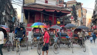 In einer belebten Straße in Kathmandu warten mehrere Rikschafahrer mit bunten Regenschirmen auf Passagiere.