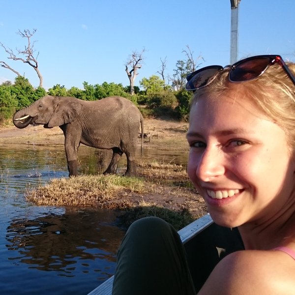 Eine junge Frau lächelt, während ein Elefant im Wasser steht, umgeben von üppigem Grün und einem klaren blauen Himmel.