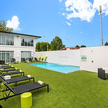 Ein einladender Poolbereich mit Liegen und grüner Rasenfläche, umgeben von einem modernen Hotelgebäude und strahlend blauem Himmel.