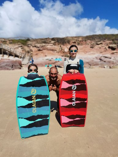 Drei Personen stehen am Strand, halten bunte Bodyboards und lächeln fröhlich in die Kamera, umgeben von einer schönen Landschaft.