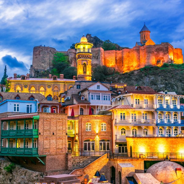 Die malerische Altstadt von Tiflis zeigt bunte Häuser und eine beeindruckende Festung im Hintergrund, beleuchtet bei Dämmerung.