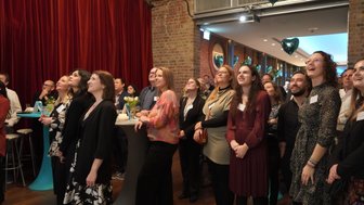 Eine Gruppe von Menschen schaut gespannt nach oben, während sie in einem festlichen Raum mit roten Vorhängen stehen.