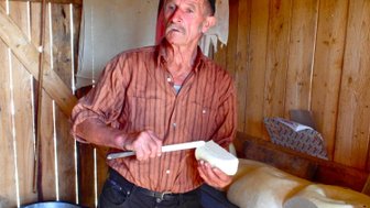 Ein älterer Mann in einem Holzraum hält ein Stück Käse und schaut überrascht, während er mit einem Messer arbeitet.