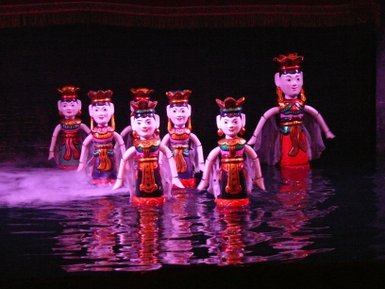 Sechs farbenfrohe Wasserpuppen stehen im Wasser, umgeben von Nebel und sanftem Licht, das die Szene mystisch erscheinen lässt.