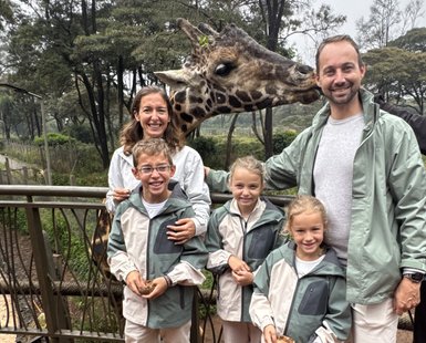 Eine Familie steht auf einer Holzterrasse, während eine Giraffe neugierig in die Kamera schaut und die Kinder lächeln.