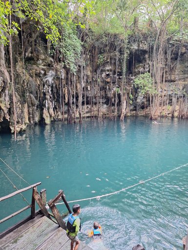 Zwei Kinder stehen an einem Holzsteg, während eines im klaren Wasser eines Cenotes schwimmt, umgeben von üppigem Grün.