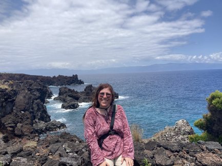 Eine Frau mit langen braunen Haaren sitzt auf einem Felsen und lächelt, während sie den Ozean und die Küste betrachtet.