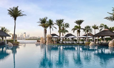 Ein ruhiger Poolbereich mit Palmen und Liegen, der die Skyline von Dubai im Hintergrund spiegelt.