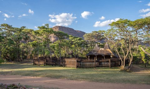 Eine rustikale Lodge mit Strohdach steht umgeben von üppigem Grün und sanften Hügeln unter einem klaren blauen Himmel.