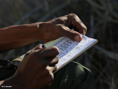 Eine Person hält ein Notizbuch und macht Notizen, während sie in einer natürlichen Umgebung sitzt.