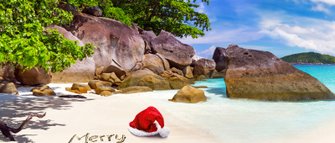 Ein roter Weihnachtsmann-Hut liegt am Strand, während 'Frohe Weihnachten' im Sand geschrieben steht, umgeben von Felsen und klarem Wasser.