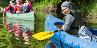 Ein fröhliches Kind paddelt in einem blauen Kajak, während andere Kajakfahrer im Hintergrund auf einem ruhigen Gewässer fahren.
