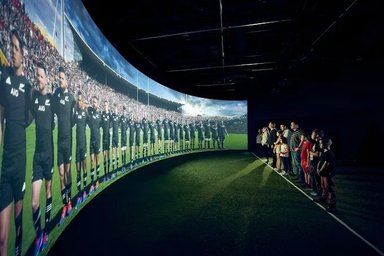 Eine Gruppe von Menschen steht vor einer großen, gebogenen Leinwand, die die All Blacks Rugby-Mannschaft zeigt.