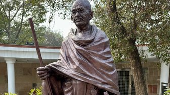 Eine bronzene Statue von Mahatma Gandhi steht auf einem Sockel, umgeben von Pflanzen und einem gepflasterten Weg.