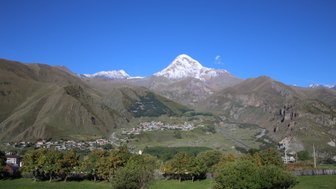Ein majestätischer Berg mit schneebedecktem Gipfel erhebt sich über eine grüne Tallandschaft und ein kleines Dorf.