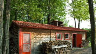 Eine rustikale Holzhütte mit rotem Dach und Fensterrahmen, umgeben von üppigem Grün und Holzstapeln.