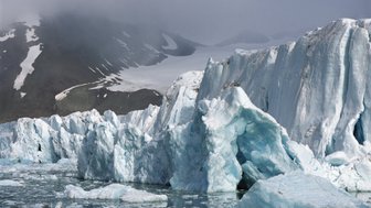 Ein beeindruckender Gletscher mit blauen Eisformationen, umgeben von schneebedeckten Bergen und einem nebligen Himmel.