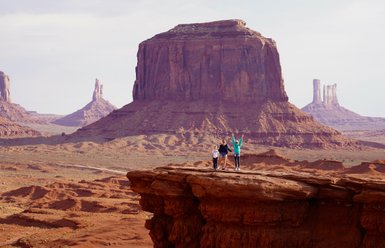 Drei Personen stehen auf einer Felsplattform in Monument Valley, umgeben von beeindruckenden roten Felsen und Hügeln.