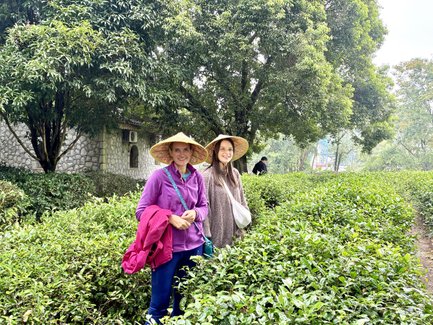 Zwei Frauen stehen in einer Teepflanzung, umgeben von üppigem Grün, beide tragen traditionelle Strohhüte.