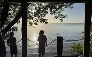 Zwei Kinder stehen am Ufer und blicken auf das ruhige Wasser, während die Sonne sanft über dem Horizont aufgeht.
