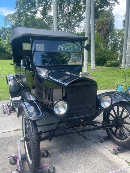 Ein klassisches schwarzes Auto aus den 1920er Jahren steht auf einem gepflasterten Weg, umgeben von üppigem Grün.
