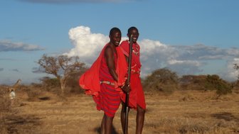 Zwei fröhliche Maasai-Krieger in leuchtend roten Gewändern posieren in der afrikanischen Savanne unter einem blauen Himmel.