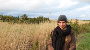 Eine lächelnde Frau steht in einem weiten Feld mit hohem Gras, umgeben von Bäumen und einem bewölkten Himmel.