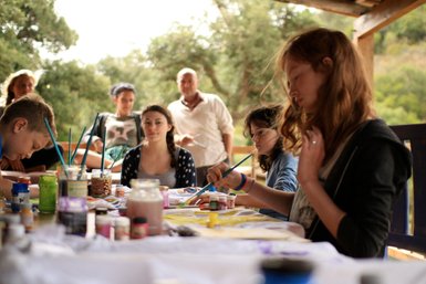 Eine Gruppe von Menschen sitzt an einem Tisch und malt mit verschiedenen Farben und Pinseln, umgeben von Natur.