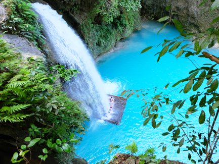 Ein Wasserfall stürzt in ein leuchtend blaues Wasserbecken, umgeben von üppigem Grün und tropischen Pflanzen.