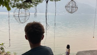 Ein Junge sitzt am Ufer des Phewa-Sees und beobachtet ein Mädchen, das am Wasser spielt, umgeben von grünen Bäumen.